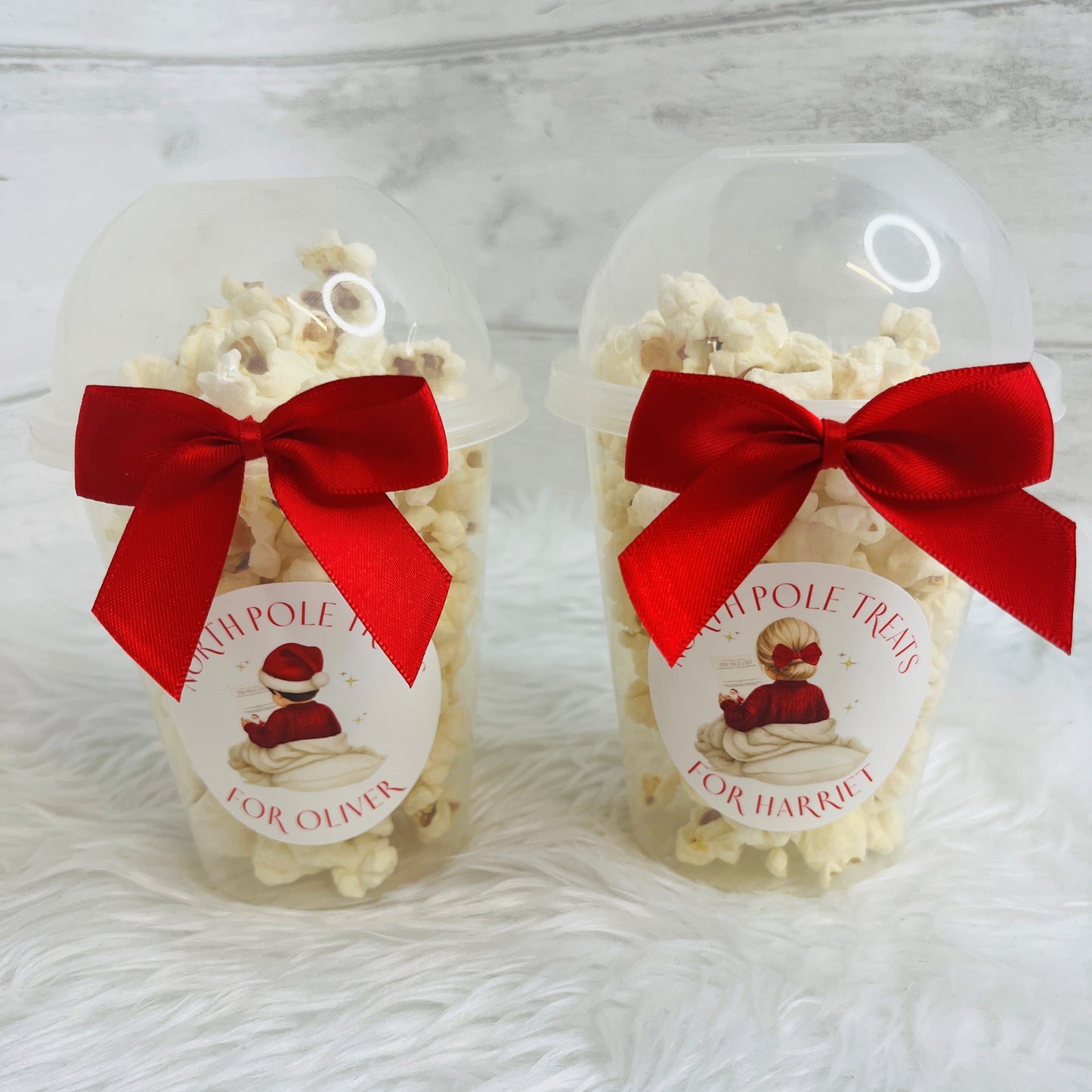 Two clear plastic popcorn cups filled with white popcorn, each tied with a large red satin bow and a round white label featuring a child in a red jumper. The labels read 'NORTH POLE TREATS' and 'FOR OLIVIER' / 'FOR HARRIET'.