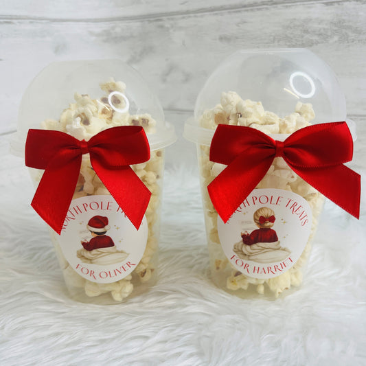 Two clear plastic popcorn cups filled with white popcorn, each tied with a large red satin bow and a round white label featuring a child in a red jumper. The labels read 'NORTH POLE TREATS' and 'FOR OLIVIER' / 'FOR HARRIET'.