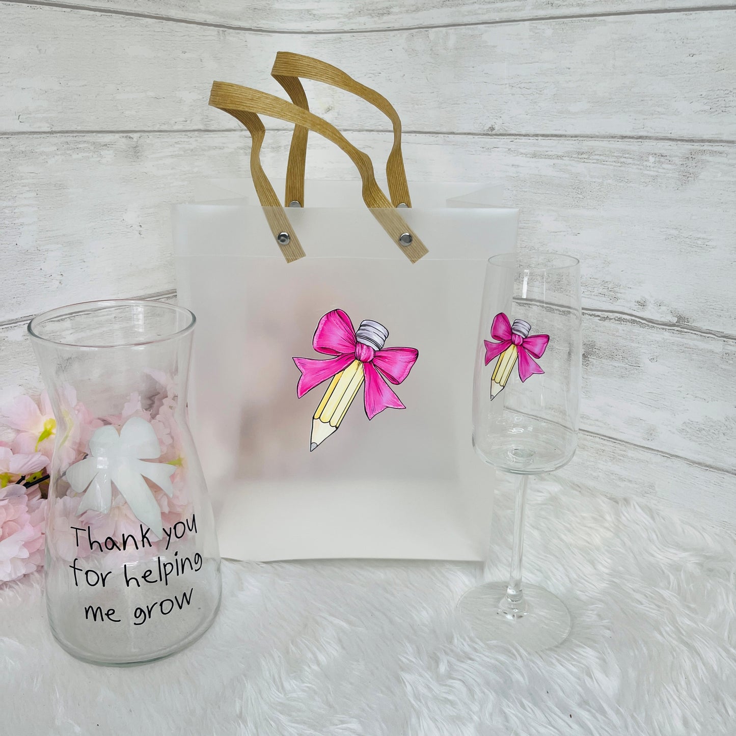 Gift bag with pink bow design, glass with 'Thank you for helping me grow' text, and champagne flute on a light surface.