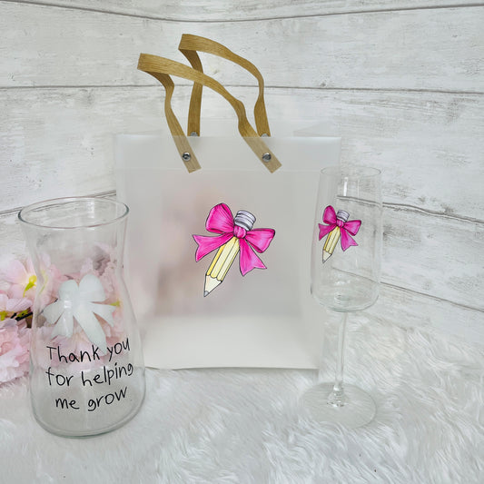 Gift bag with pink bow design, glass with 'Thank you for helping me grow' text, and champagne flute on a light surface.
