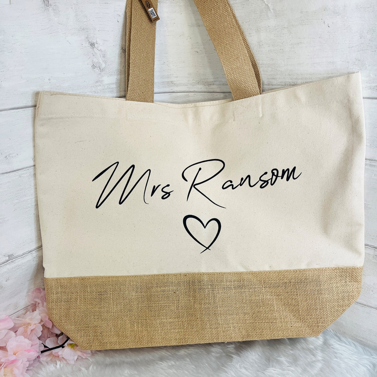 A canvas tote bag with a natural cream upper section, natural tan burlap lower section, and tan shoulder straps. The front is printed with the black cursive text 'Mrs Ransom' and a black heart outline.