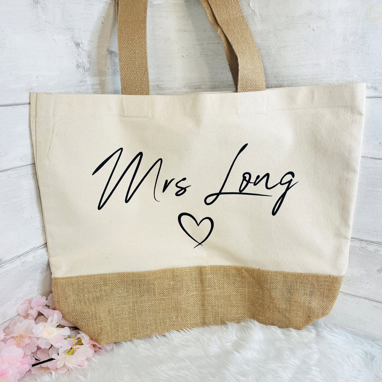 A canvas tote bag with a natural burlap lower section and handles, featuring the words 'Mrs Long' and a heart in black script on the front, styled on a white faux fur surface against a white wood plank background.