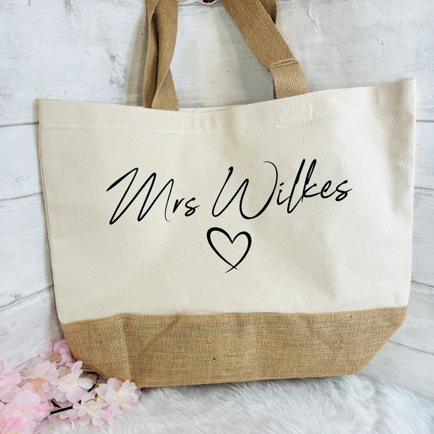 A canvas tote bag with a natural burlap bottom panel and matching handles, printed with the text 'Mrs Wilkes' and a heart in black script on the front, styled on a white faux fur surface with pink flowers against a white wood background.