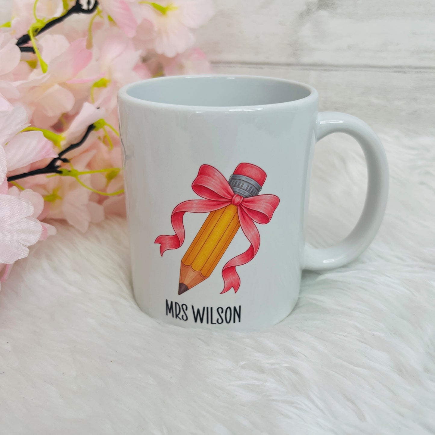 A white ceramic mug with a printed illustration of a yellow pencil tied with a pink ribbon, above the text 'MRS WILSON', set on a white faux-fur surface with pink flowers in the background.