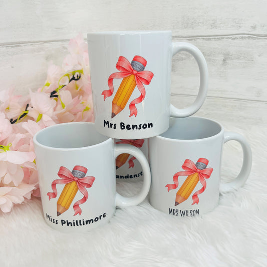 Four white ceramic mugs decorated with a yellow pencil and pink ribbon illustration, displayed on a white faux-fur surface with pink flowers and a light wood background.