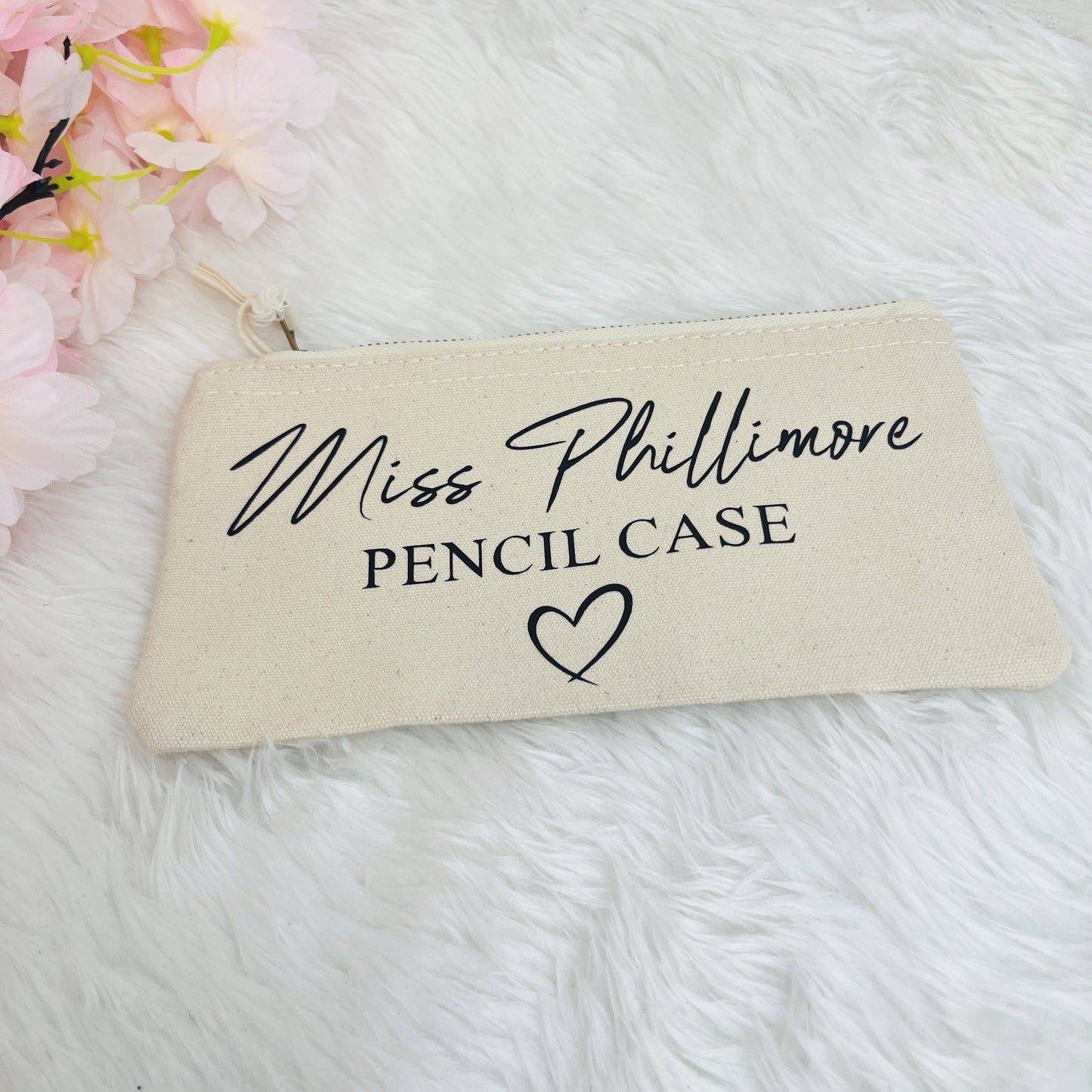 A cream-colored canvas pencil case with black printed text reading 'Miss Phillimore PENCIL CASE' and a heart, displayed on a white faux fur surface with pink flowers in the corner.