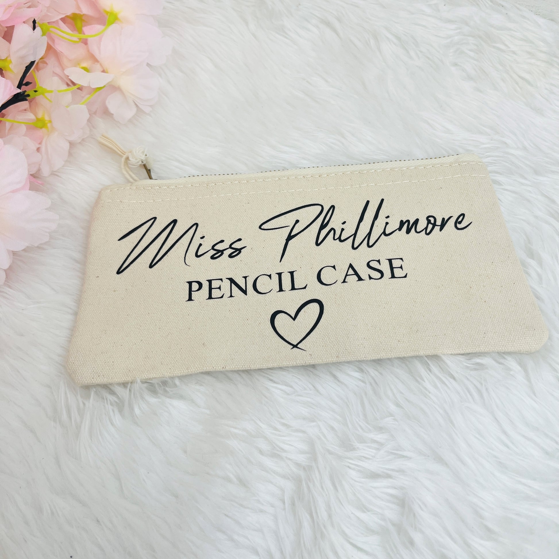 A cream-colored canvas pencil case with black printed text reading 'Miss Phillimore PENCIL CASE' and a heart, displayed on a white faux fur surface with pink flowers in the corner.