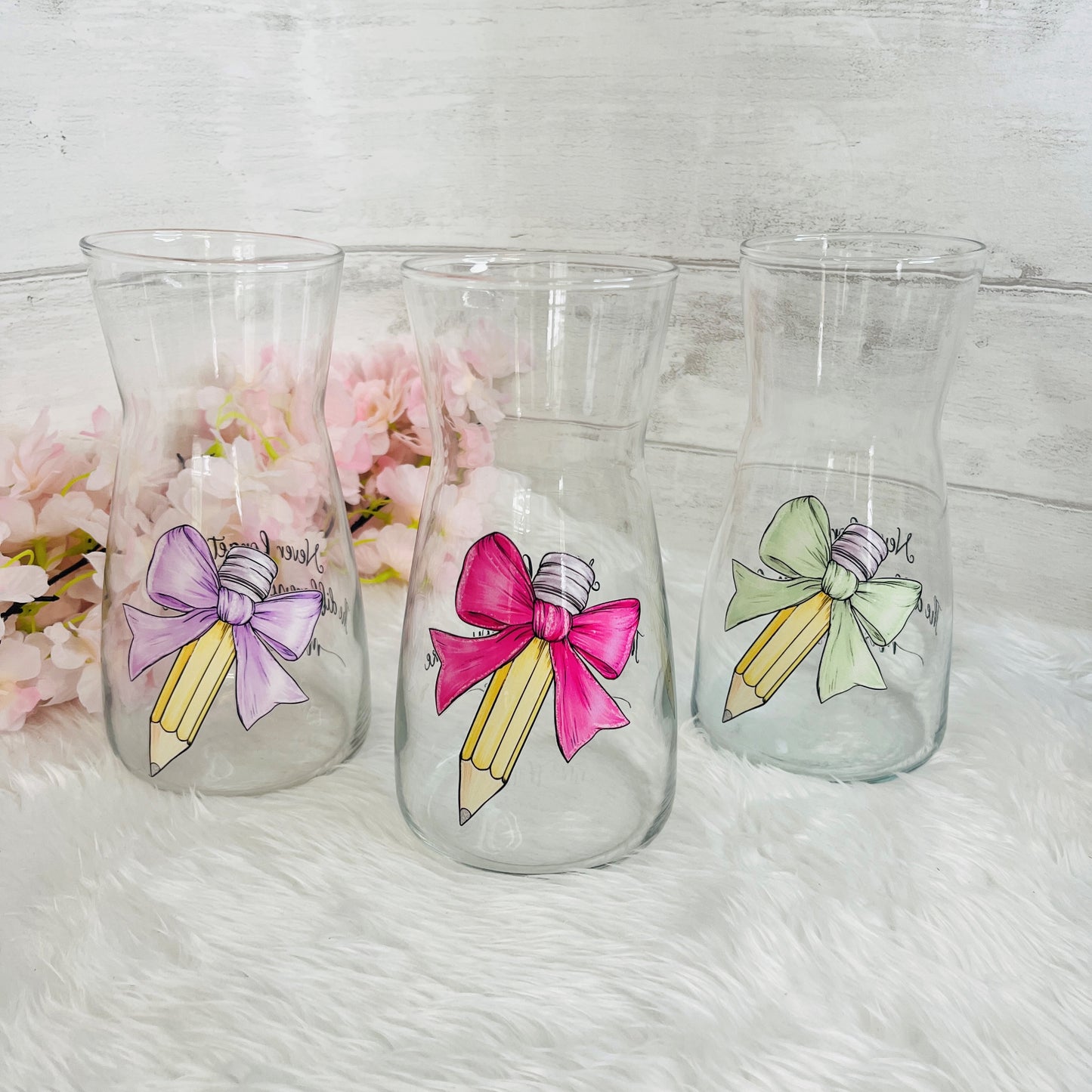 Three clear glass vases decorated with  pencil and bow designs in purple, pink, and green, displayed on a white faux fur surface with pink flowers in the background.