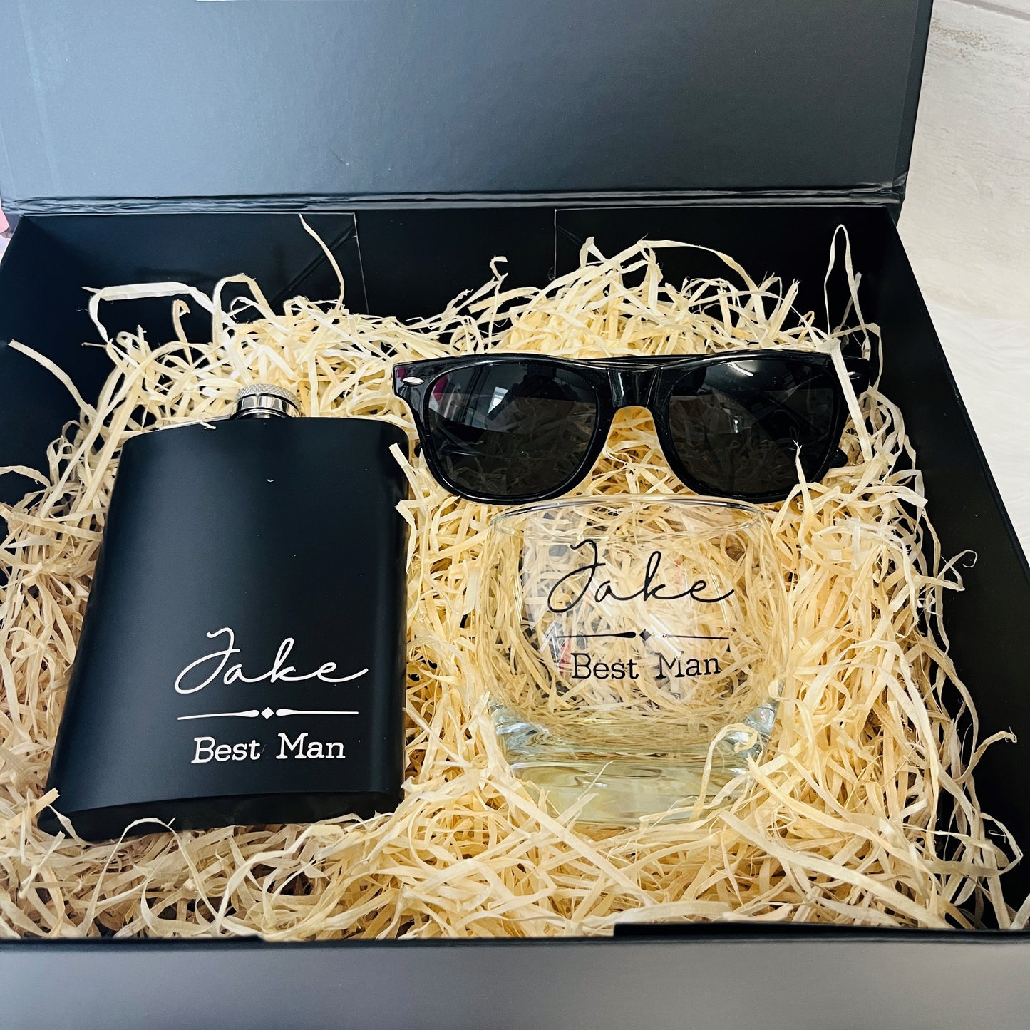 Black flask, glass with 'Best Man' text, and sunglasses in a box with straw bedding.