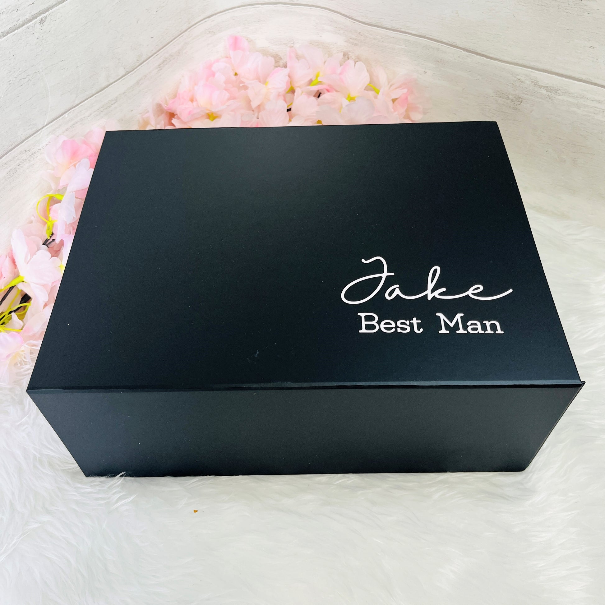 Black box with 'Jake Best Man' text on a white surface with pink flowers.