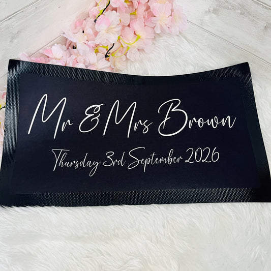 A black rectangular beer mat personalised with white script text reading 'Mr & Mrs Brown' and 'Thursday 3rd September 2026', styled on a white faux-fur surface with pink flowers in the background.