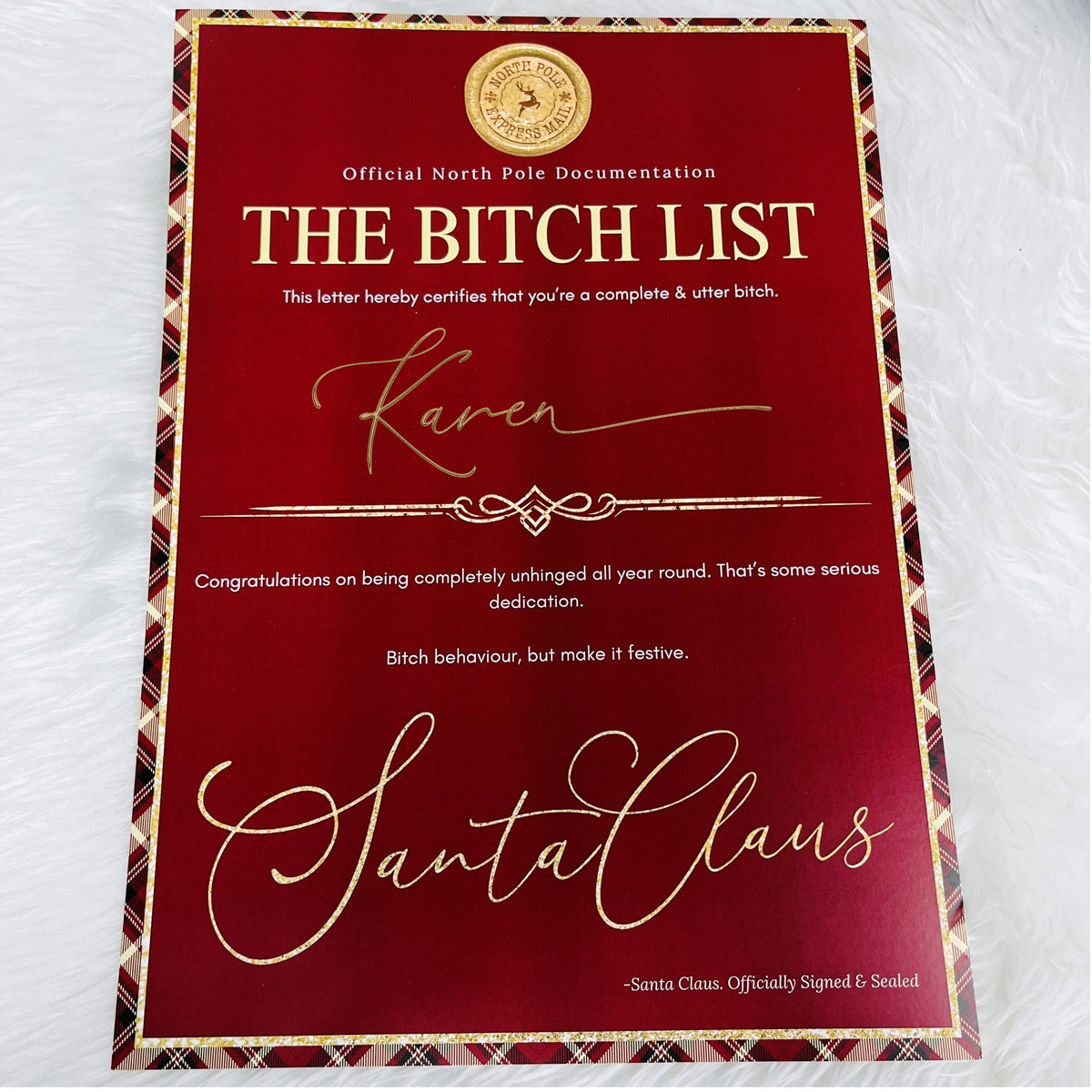 A red and gold novelty letter certificate titled 'THE BITCH LIST' with decorative gold script and text, photographed flat on a white textured fabric background.