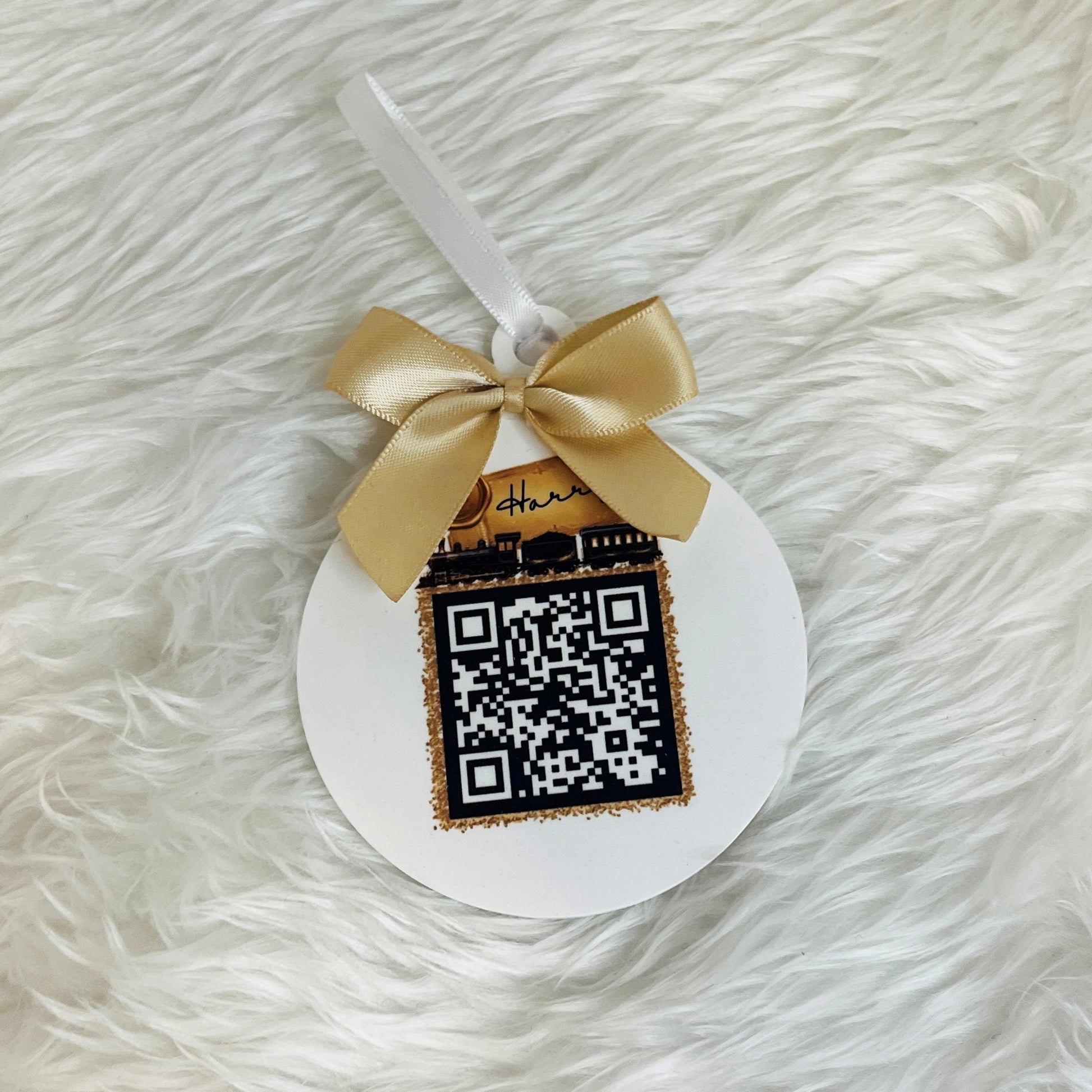 A round acrylic white santa tracker ornament with a white ribbon loop, decorated with a gold satin bow and a black-and-white QR code panel, placed on a white faux fur background.