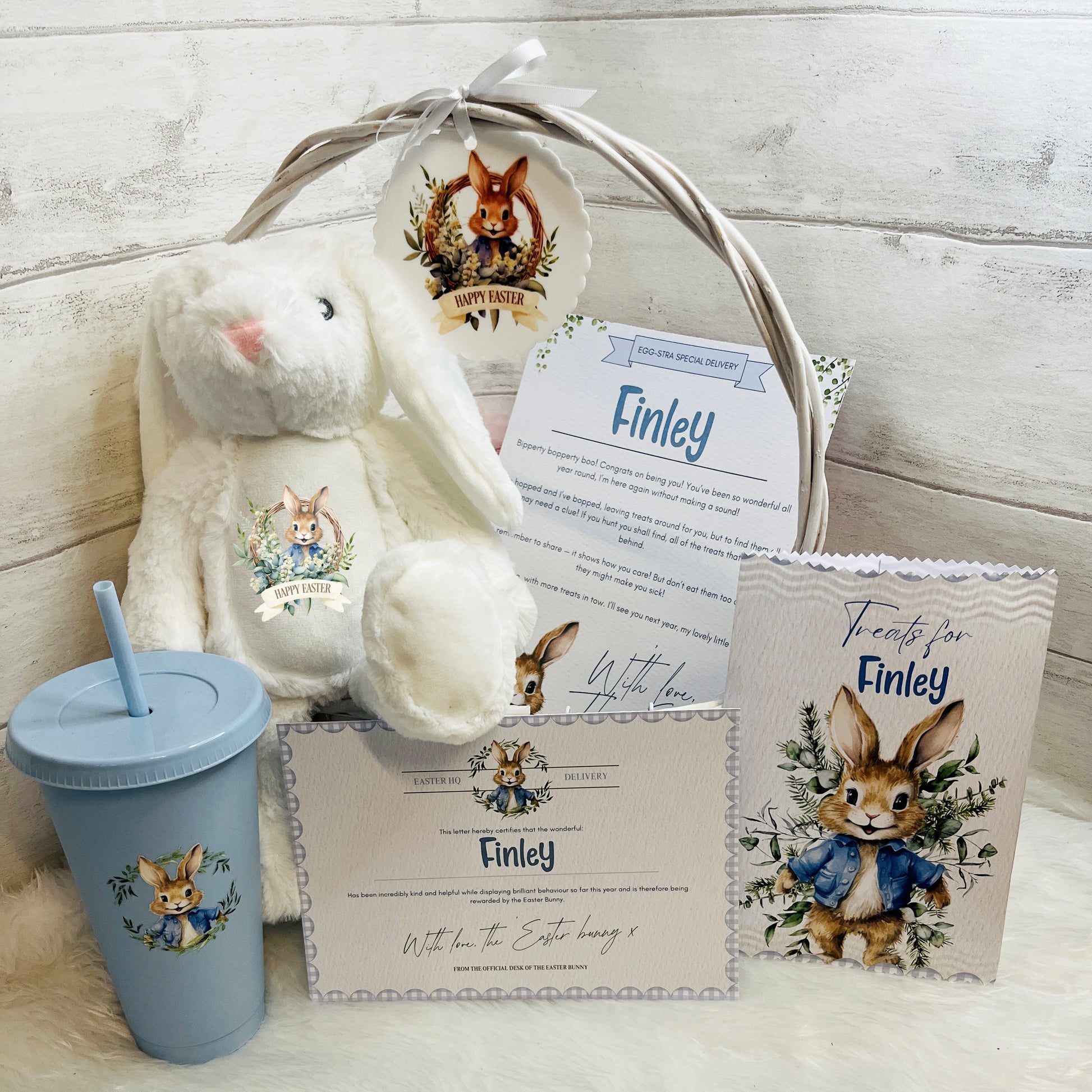 Blue Easter gift basket with plush bunny, tumbler, and personalized cards on a white fluffy surface.