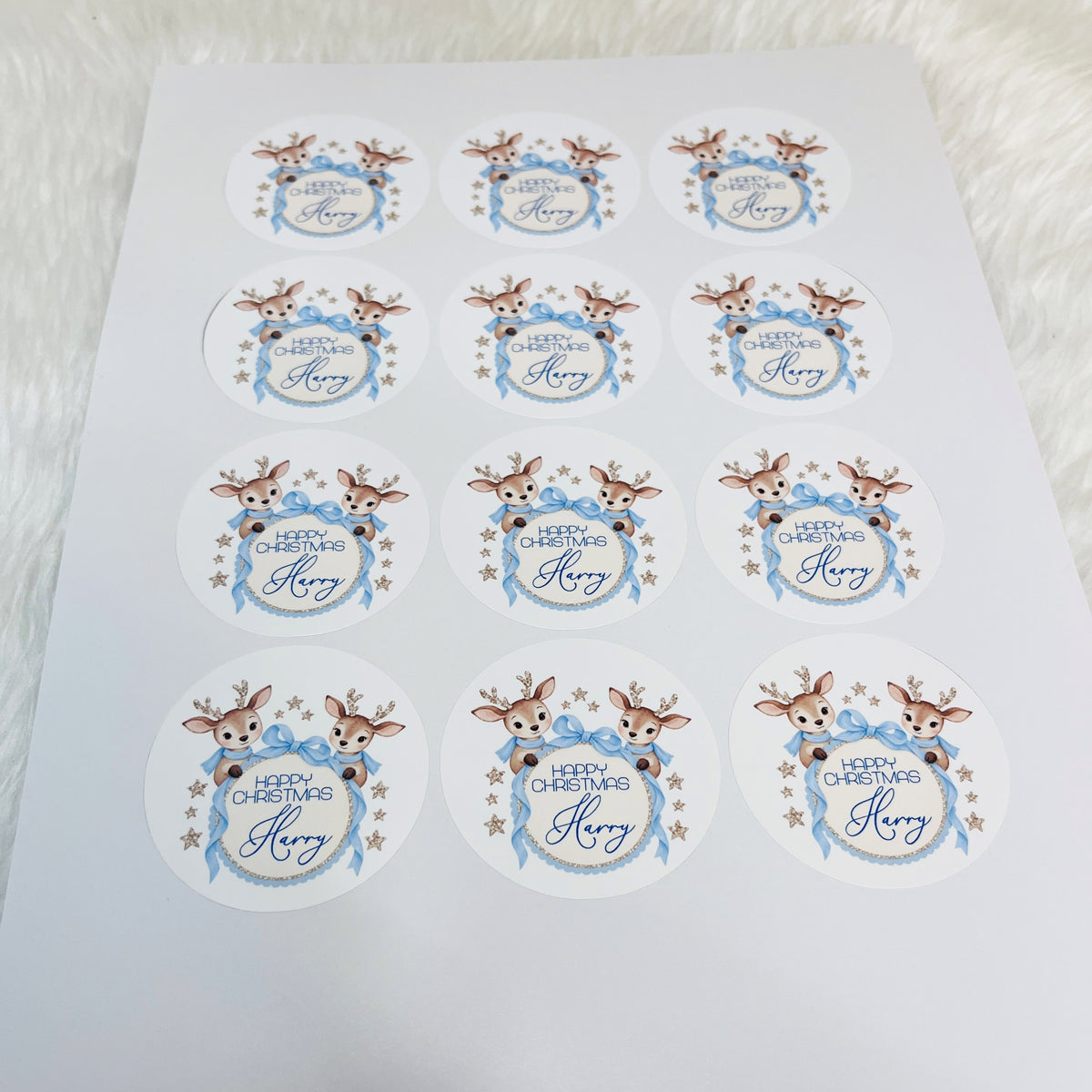 A sheet of round white Christmas stickers featuring illustrated brown reindeer with antlers, blue bows and ribbons, gold stars, and the text 'HAPPY CHRISTMAS Harry' on a white background.