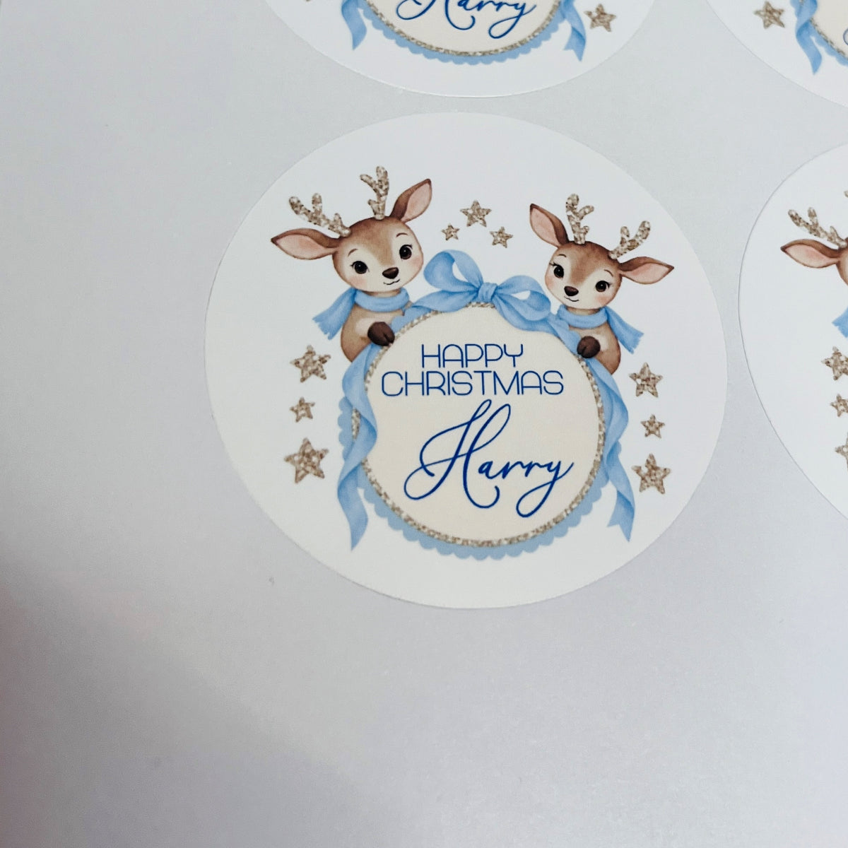 A round white Christmas sticker featuring two cartoon deer with antlers, blue bows, gold stars, and the text 'Happy Christmas Harry' in blue script, shown on a light gray background with other partial stickers visible around the edges.