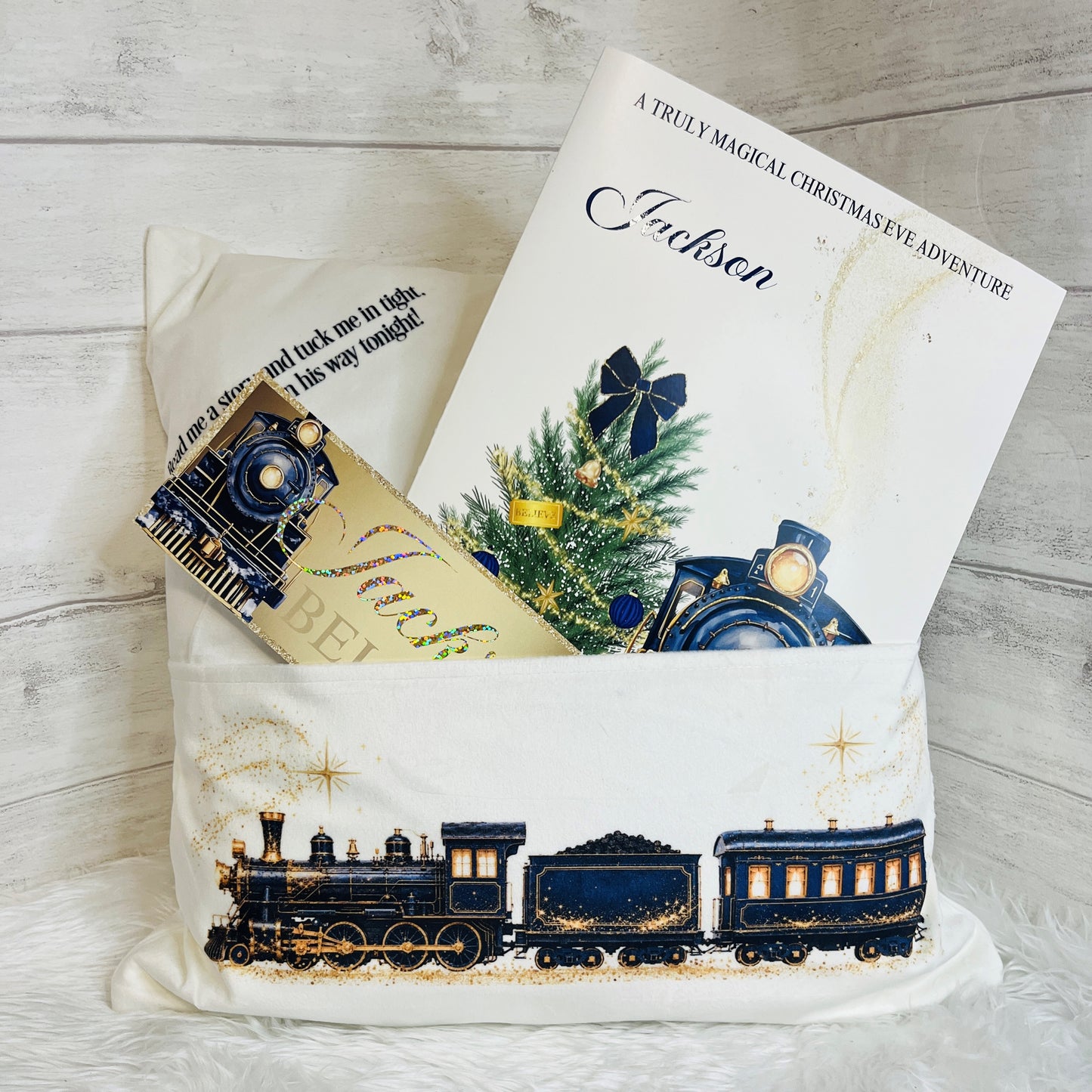 A navy and gold Christmas gift pillow featuring a vintage-style navy train illustration on the front, styled with a personalised Christmas story book  and a small gold ticket on a white faux fur surface against a light wood plank background.