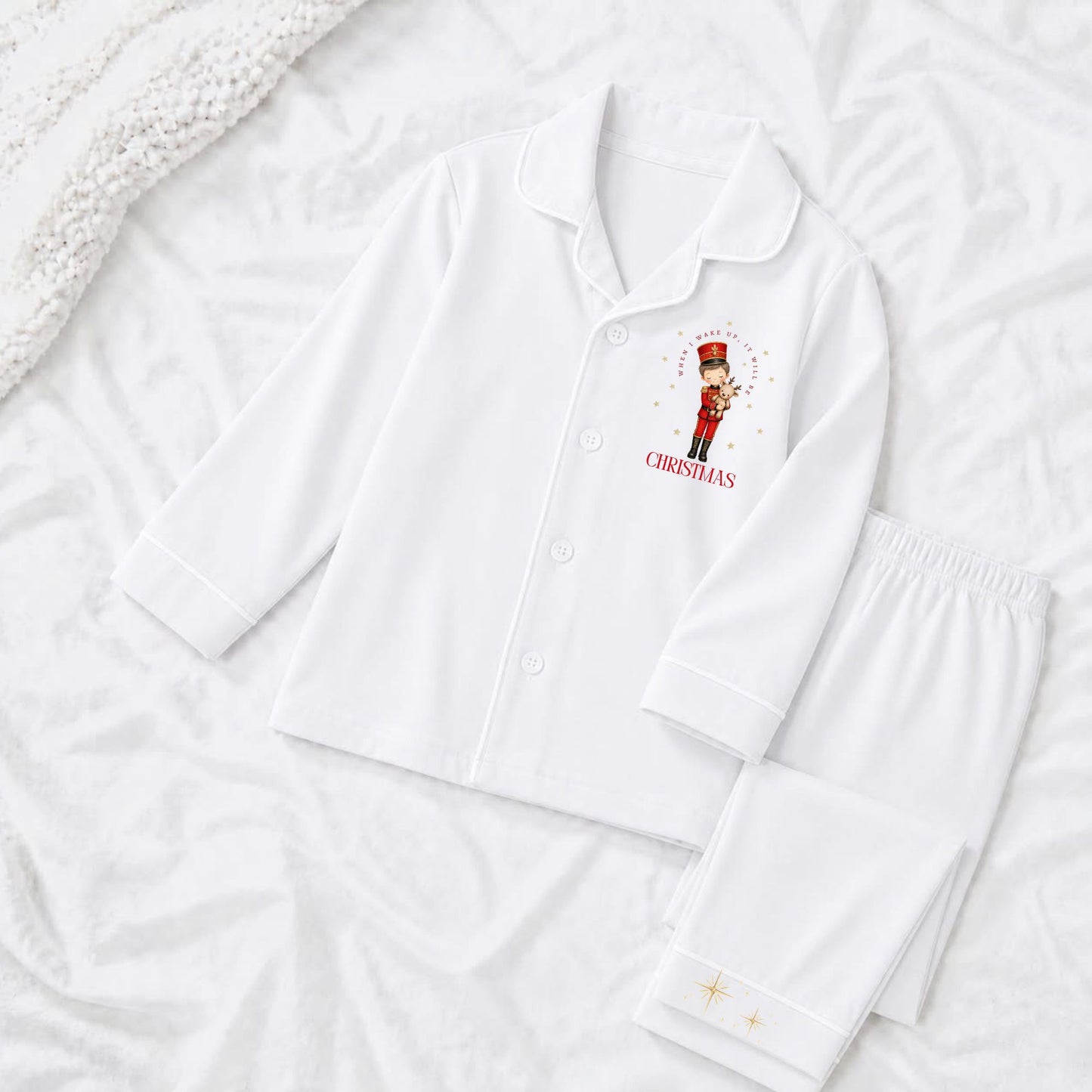 A white long-sleeve pajama top and matching pants laid flat on a white textured blanket. The pajama top has a small red and gold nutcracker graphic with the word 'when i wake up it will be christmas' on the left chest.