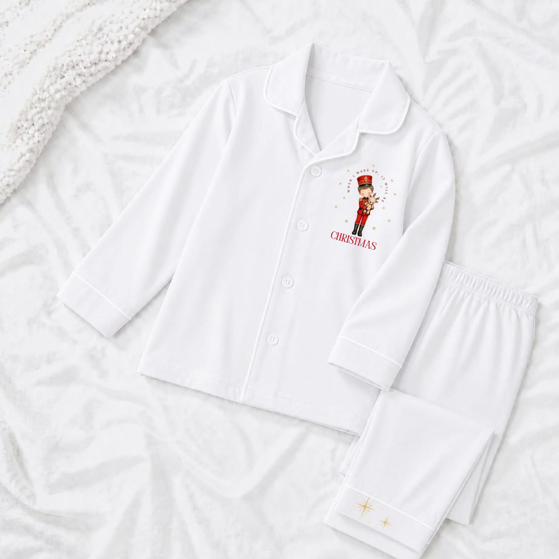 A white long-sleeve pajama top and matching pants laid flat on a white textured blanket. The pajama top has a small red and gold nutcracker graphic with the word 'when i wake up it will be christmas' on the left chest.