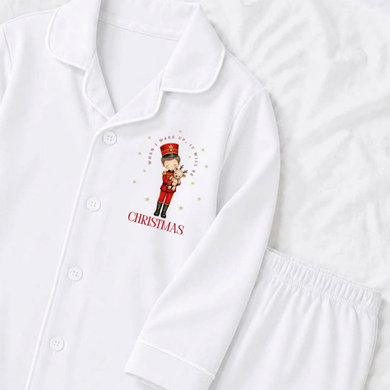 White long-sleeve pajama top with a button front and collar, featuring a Christmas nutcracker graphic with the word 'when i wake up it will be christmas' on the chest, shown on a white textured background with matching white pajama trousers partially visible.