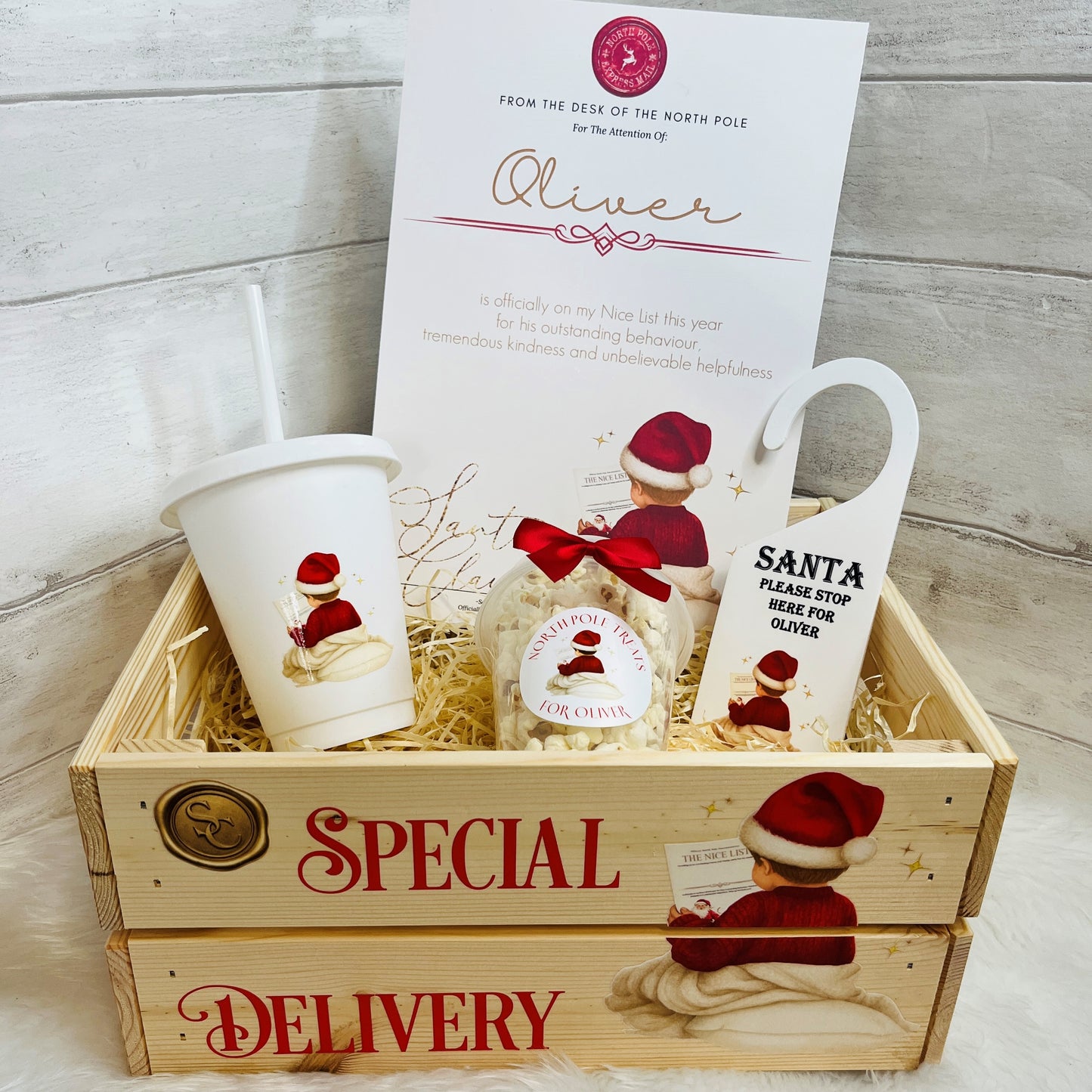 A Christmas-themed gift basket set in a wooden crate with 'SPECIAL DELIVERY' text. The set includes a white Santa cup, a snack pot of Christmas treats, a white Santa Door hanger, and a white A4 nice list certificate, all arranged in shredded paper inside a light wood crate.