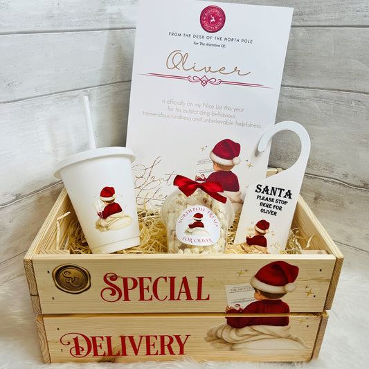 A Christmas-themed gift basket set in a wooden crate with 'SPECIAL DELIVERY' text. The set includes a white Santa cup, a snack pot of Christmas treats, a white Santa Door hanger, and a white A4 nice list certificate, all arranged in shredded paper inside a light wood crate.