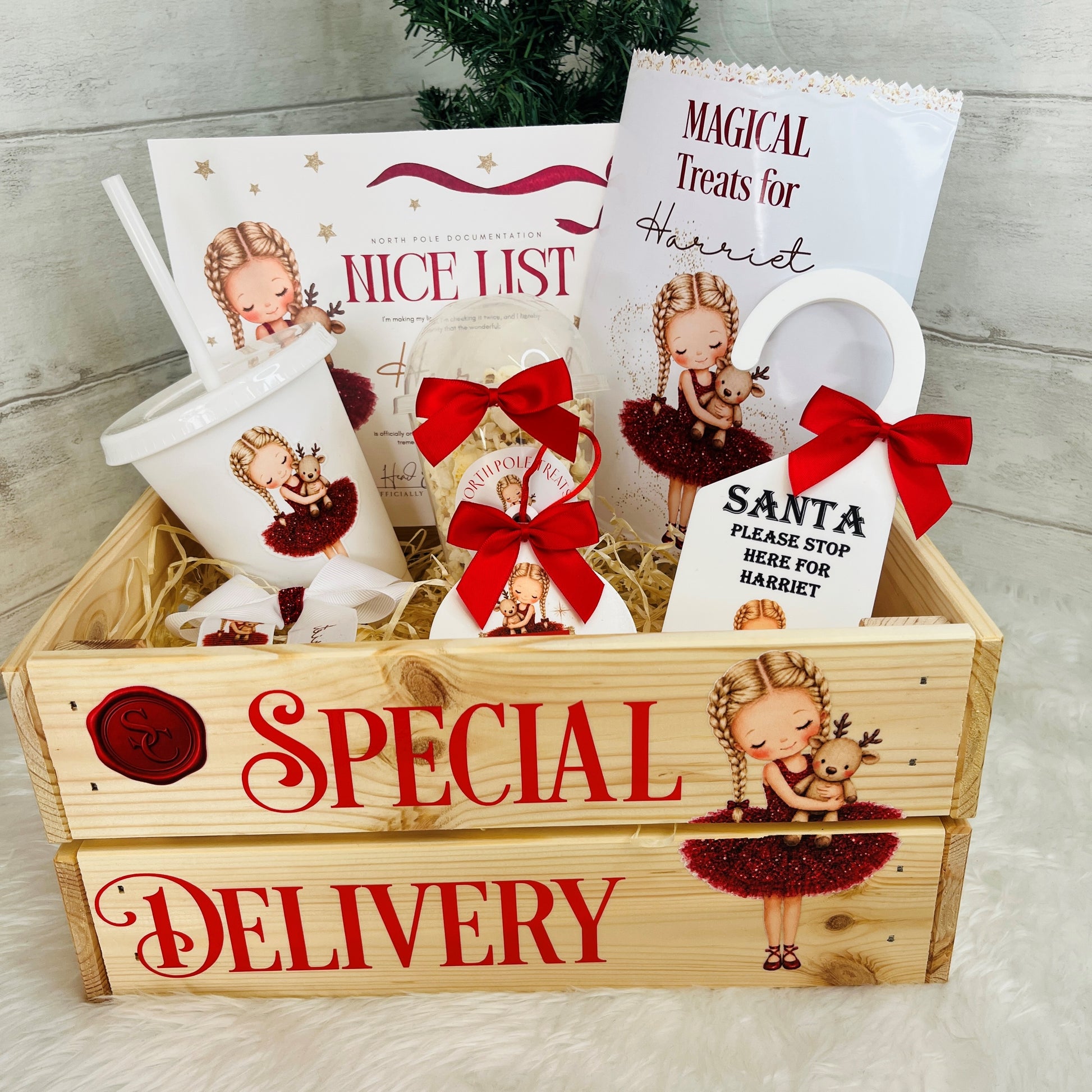 A wooden crate gift box labeled 'SPECIAL DELIVERY' filled with christmas-themed printed items, including a cup, Nice List, a treat bag, a snack pot, santa tracker, hair bow and door hanger on a light neutral background