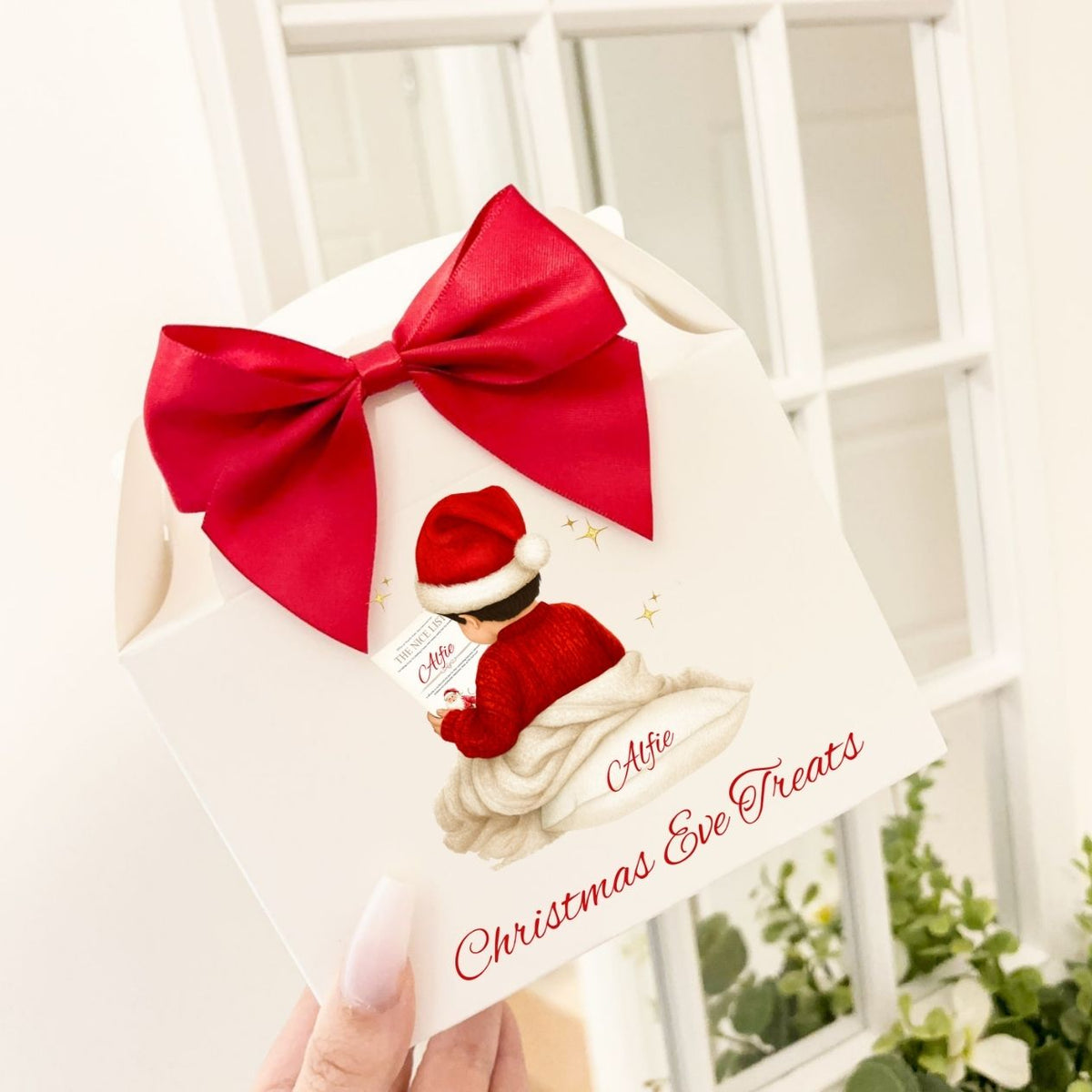 A hand holds a white Christmas Eve treats gift box decorated with a large red bow, featuring an illustration of a child in a Santa hat and the text 'Alfie Christmas Eve Treats'.
