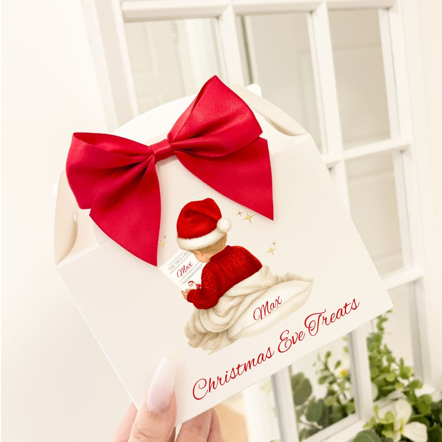 A hand holding a white Christmas Eve treat box decorated with a large red satin bow and an illustration of a young boy with blonde hair and a santa hat with a red jumper reading a 'Santa's Nice List certificate. The box has the text 'Max's Christmas Eve Treats' in red vinyl