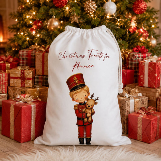 A white Christmas-themed drawstring Santa sack with a printed illustration of a child soldier nutcracker holding a teddy bear and the text 'Christmas Treats for Ronnie', displayed in front of a decorated Christmas tree with wrapped gift boxes.
