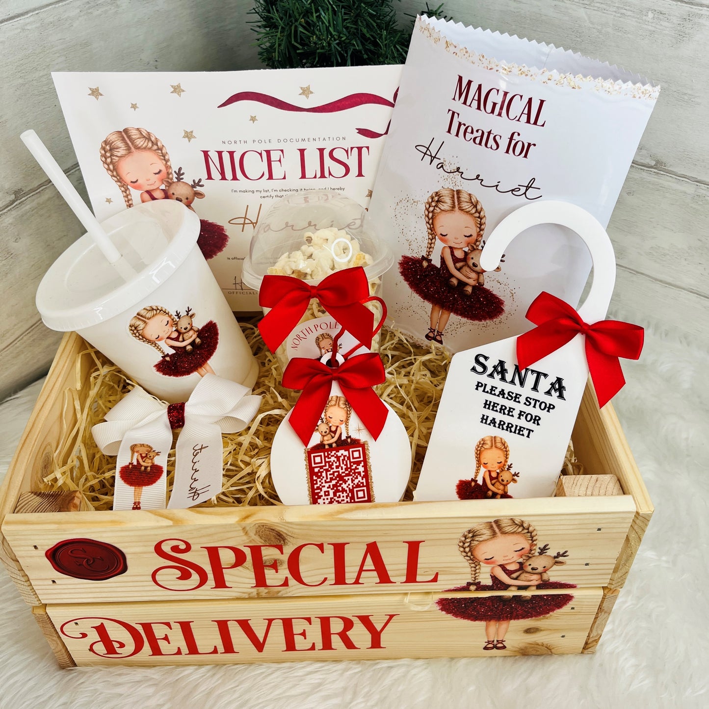 aerial photo of a wooden crate gift box labeled 'SPECIAL DELIVERY' filled with christmas-themed printed items, including a cup, Nice List, a treat bag, a snack pot, santa tracker, hair bow and door hanger on a light neutral background 