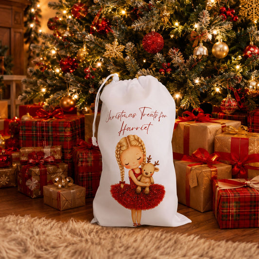 A white Christmas-themed drawstring gift sack with an illustration of a girl in a red tutu holding a reindeer, with the text 'Christmas Treats for Harriet', placed in front of a decorated Christmas tree with wrapped presents on a wooden floor.