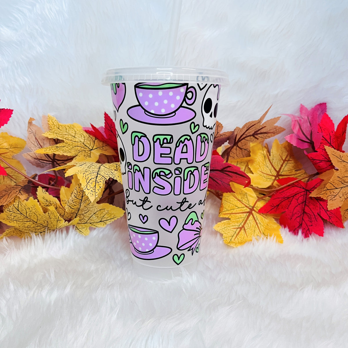 A clear plastic tumbler cup with a printed design featuring purple tea cups, a skull, hearts, and the text 'DEAD INSIDE but cute', displayed on a white faux fur surface with autumn leaves around it.