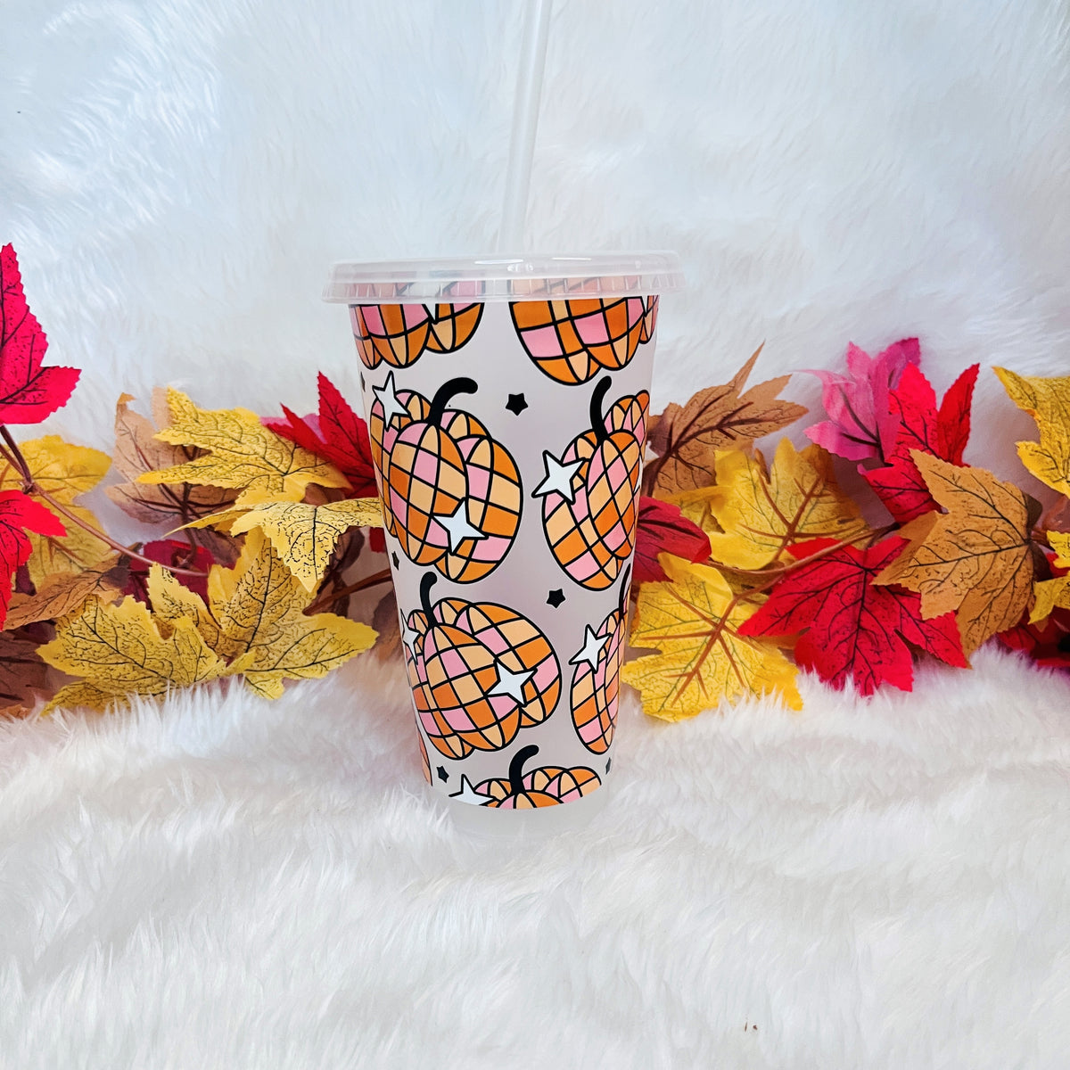 A clear plastic cup with a lid and straw, printed with a pattern of orange and yellow plaid pumpkins set on a white faux fur surface with colorful autumn leaves around it.