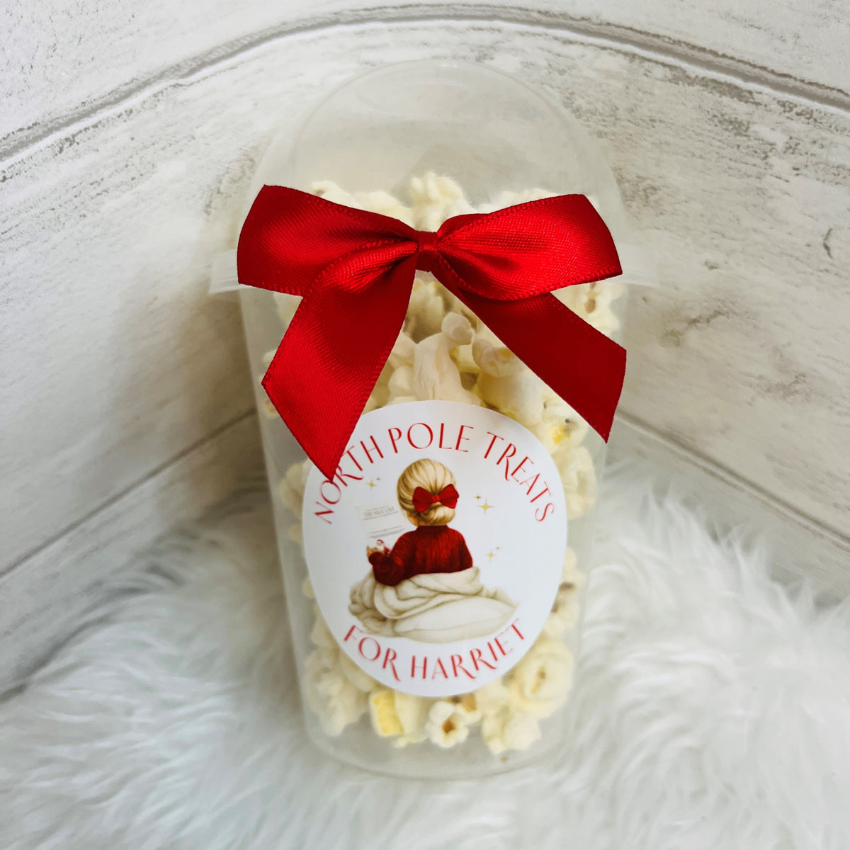A clear cylindrical container filled with popcorn and topped with a large red satin bow, featuring a round white label that reads 'North Pole Treats For Harriet.' The container is photographed on a white faux-fur surface against a distressed white wood background.