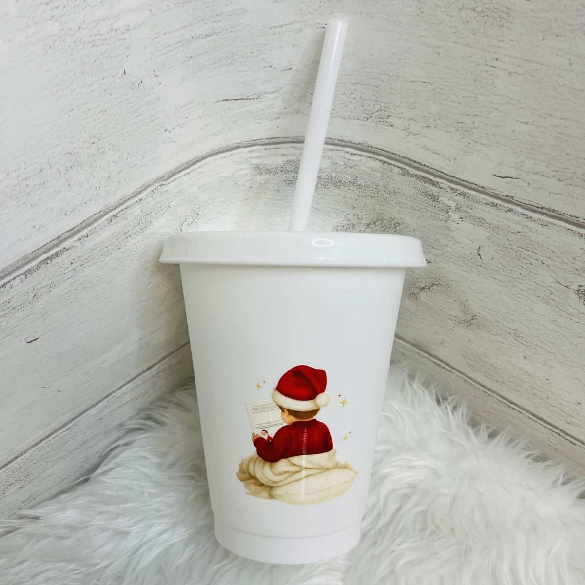 A white 16oz plastic tumbler cup with a  lid and straw, featuring a printed illustration of a child in a red Santa hat and red sweater sitting on a cream blanket and reading a letter. The cup is placed on a white faux-fur surface against a whitewashed wood background.