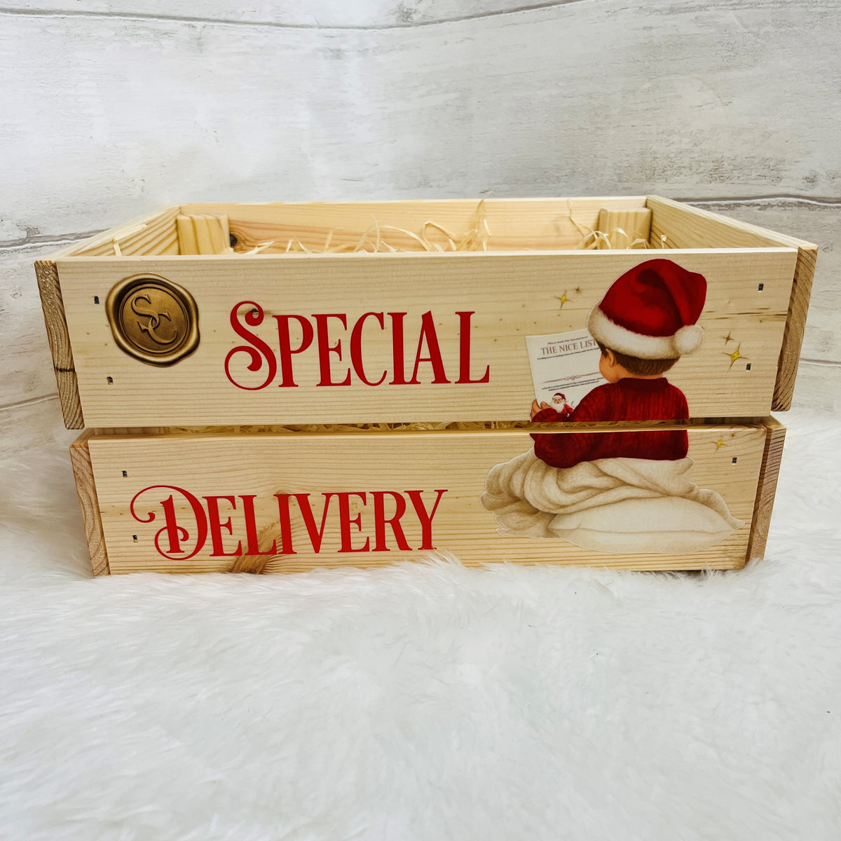 A rustic wooden crate with two stacked drawers, decorated with red text reading 'SPECIAL DELIVERY' and a Christmas illustration of a child in a Santa hat. The crate is filled with straw and sits on a white faux fur surface against a light gray wood plank background.