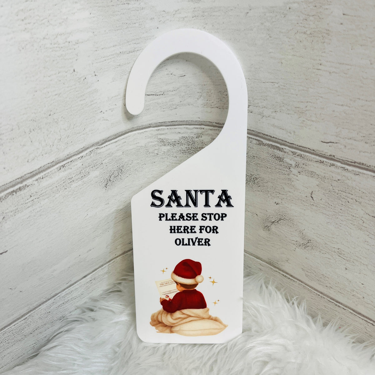 A white Acrylic Christmas door hanger with a custom printed design featuring the text 'SANTA PLEASE STOP HERE FOR OLIVER' and an illustration of a boy in a red jumper with a santa hat, set against a rustic white wood background with a white faux fur surface.