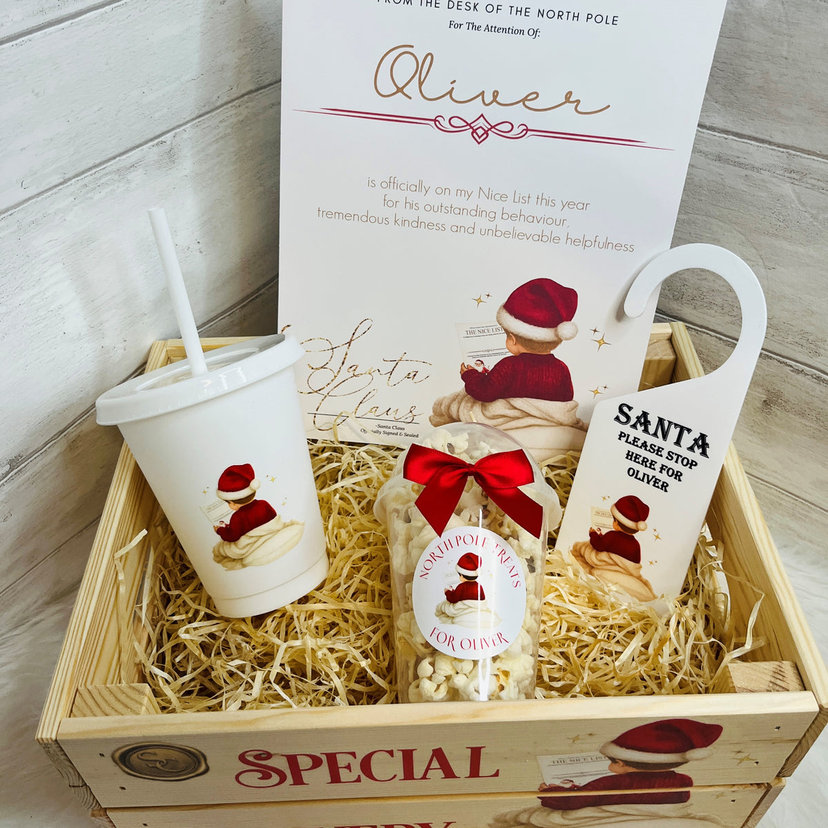 A Christmas-themed gift basket set in a wooden crate with shredded paper filler, featuring a white plastic cup with a straw, a snack pot of 'North Pole Treats', an A4 nice list kids certificate, Santa 'Please stop here for Oliver' Acrylic Door hanger.