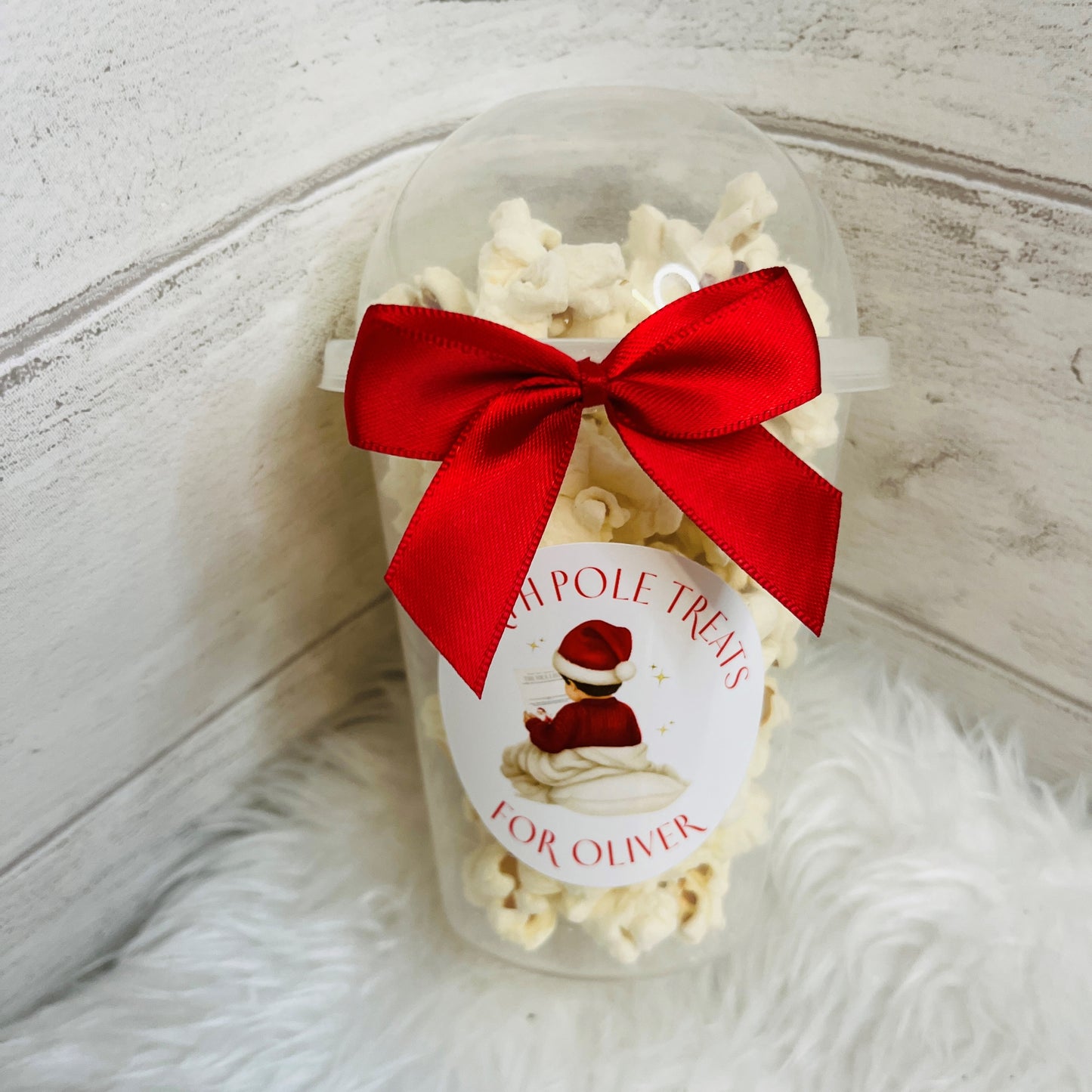 A clear plastic cup filled with white popcorn and tied with a large red ribbon bow, featuring a round white label that reads 'North Pole Treats For Oliver' with an illustration of a child in a Santa hat. The cup is placed on a white faux-fur surface against a white wood background.