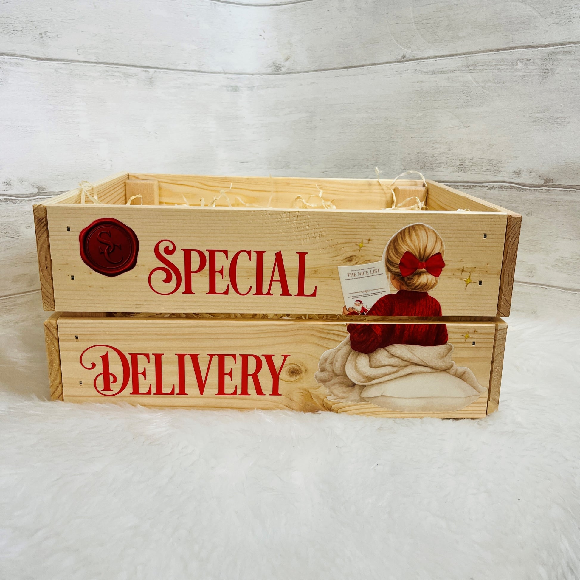 A rustic wooden crate with two stacked front panels, printed with 'SPECIAL DELIVERY' in red script, a red wax-seal style emblem, and an illustration of a girl in a red jumper and bow. The crate is photographed on a white faux-fur surface against a light gray wood-plank backdrop.