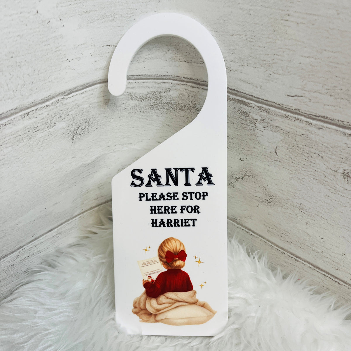 A white plastic Christmas door hanger with a custom printed design. The design features the text 'SANTA PLEASE STOP HERE FOR HARRIET' in black font above an illustration of a child in a red Jumper and hair bow.