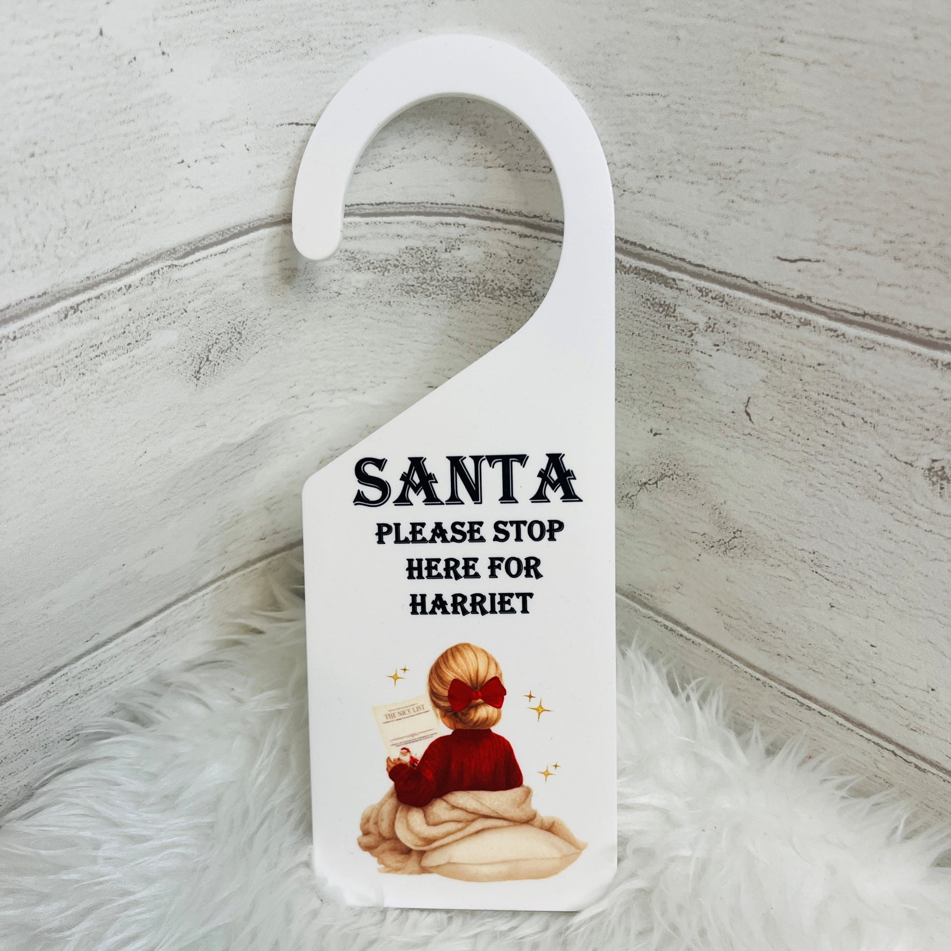 A white plastic Christmas door hanger with a custom printed design. The design features the text 'SANTA PLEASE STOP HERE FOR HARRIET' in black font above an illustration of a child in a red Jumper and hair bow.