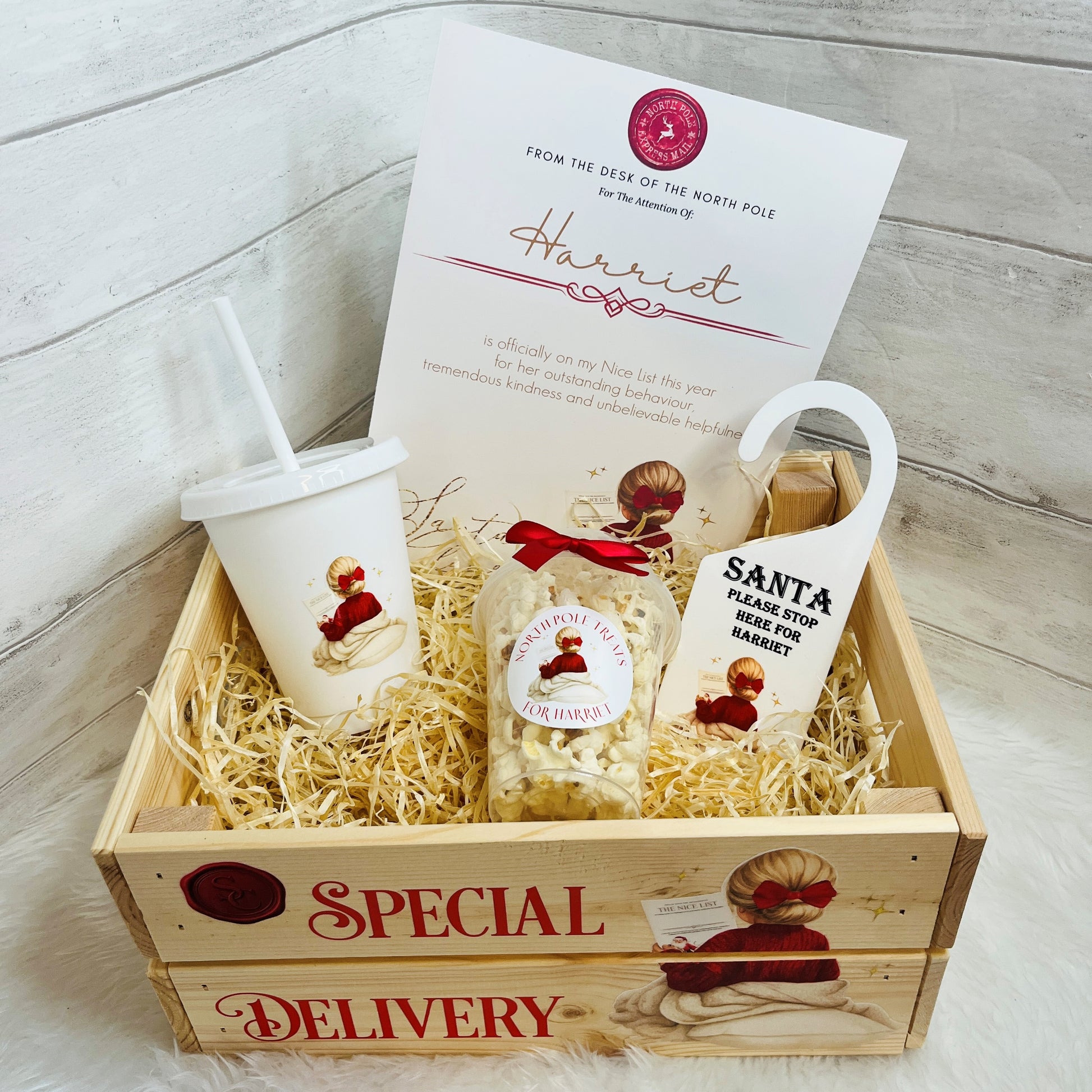 A Christmas-themed gift basket set in a light wood crate with shredded paper filler. The set includes a white cold cup with a straw, a printed A4 Nice list card addressed to 'Harriet', a snack pot of popcorn with a Santa-themed label, and a white Santa hanger tag.