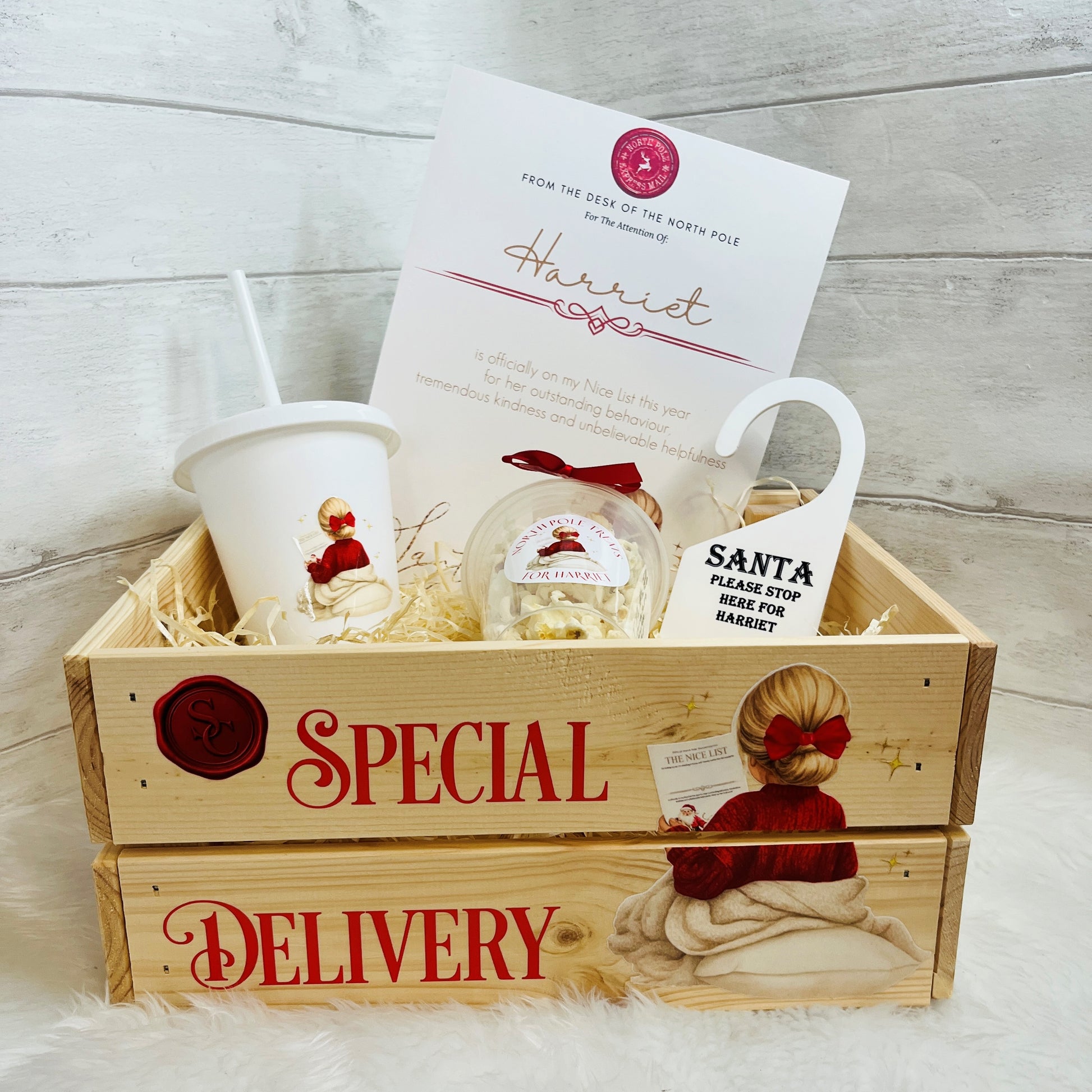 A Christmas-themed gift basket set in a wooden crate labeled 'SPECIAL DELIVERY,' containing a white Santa cup with straw, a Santa snack pot and an A4 North Pole printed card, arranged on a white faux-fur surface against a light wood plank background.