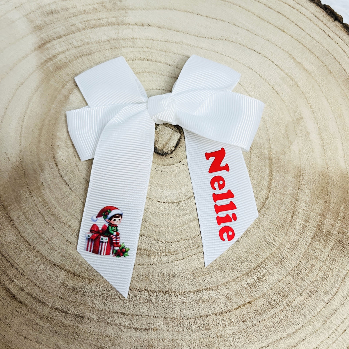 A white grosgrain ribbon hair bow personalised with the name 'Nellie' in red lettering and a small Christmas elf graphic, displayed on a natural wood slice background.