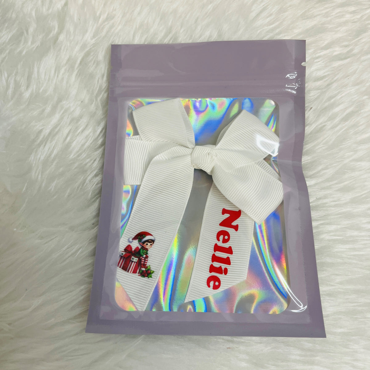A personalised white grosgrain ribbon bow with a red 'Nellie' name print and a small elf graphic, packaged inside a lavender zip-top pouch with an iridescent holographic insert, set on a white faux fur background.