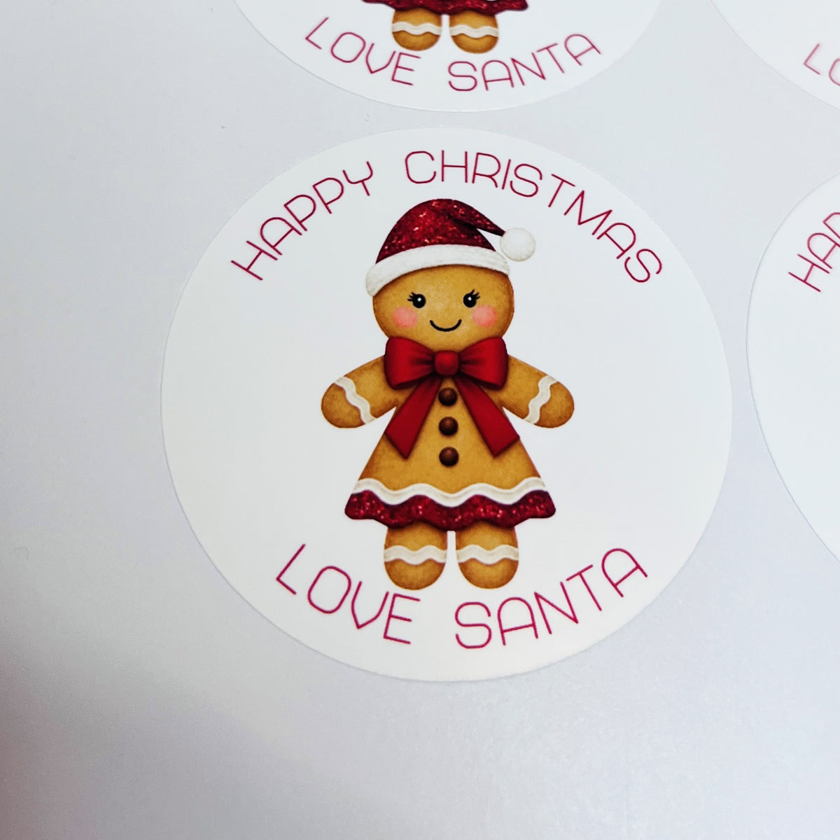 A round white sticker featuring a smiling gingerbread girl in a red Santa hat and bow tie, with the words 'HAPPY CHRISTMAS LOVE SANTA' in red around the illustration.