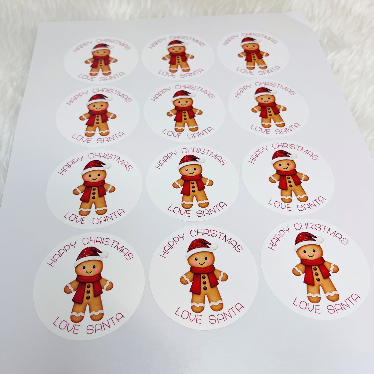 A sheet of round white stickers featuring a cute gingerbread man illustration and the text 'HAPPY CHRISTMAS LOVE SANTA' in red, laid out on a white textured background.