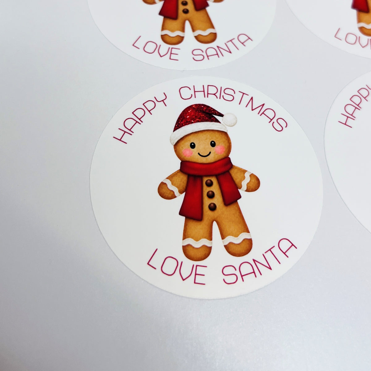 Round white Christmas-themed stickers featuring a gingerbread man wearing a red Santa hat and scarf, with the text 'HAPPY CHRISTMAS LOVE SANTA' in red, shown on a light gray background.